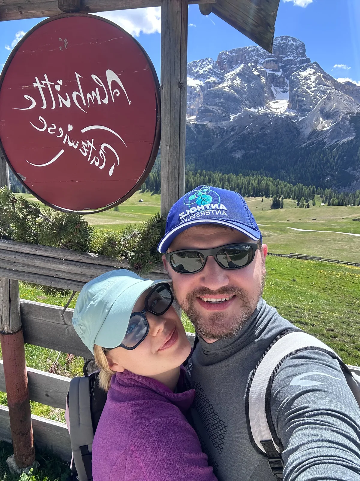 Velena and Dragos beside the Almhuette Edelweiss sign at Prato Piazza, a Dolomite peak rising behind the alpine meadow